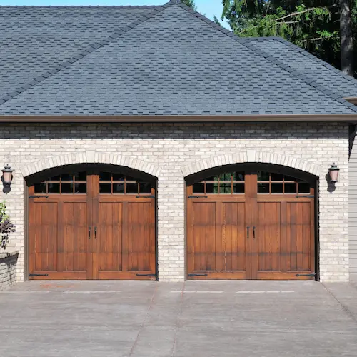 Front exterior of a home with two side-by-side arched wooden garage doors featuring windows and decorative hardware